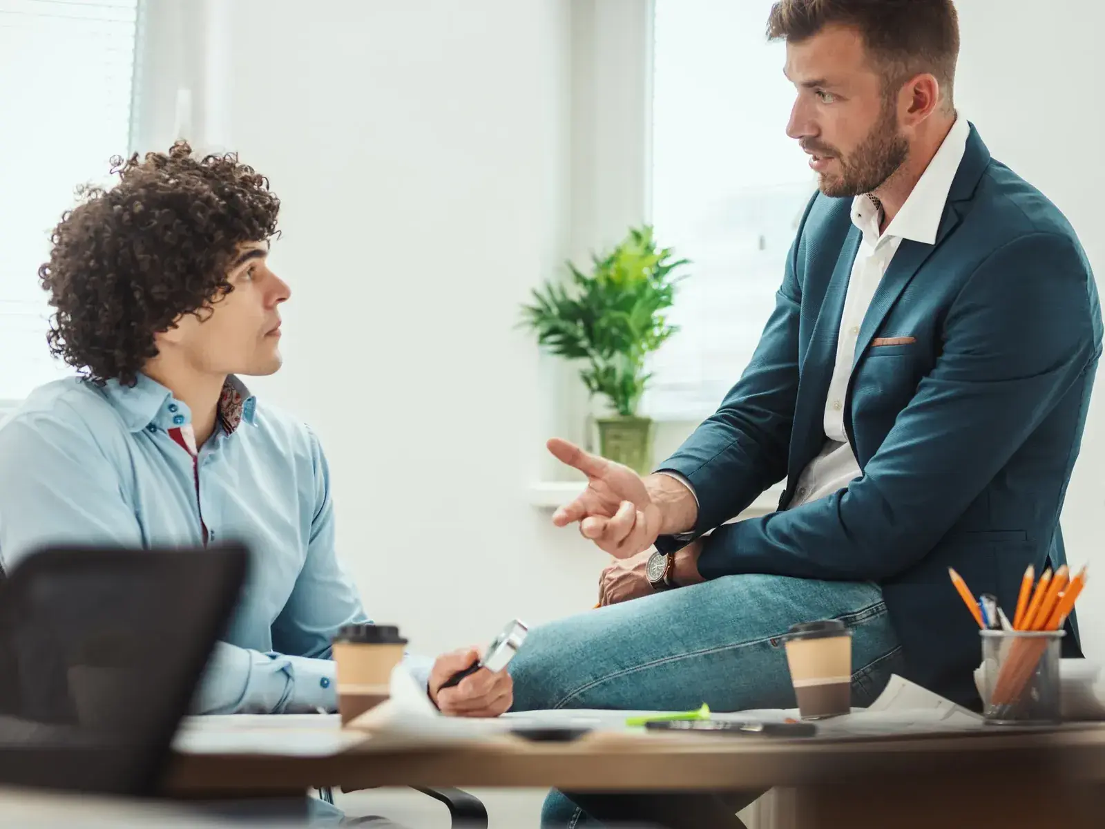 Een manager zit op de rand van het bureau en legt een medewerker uit hoe het moet