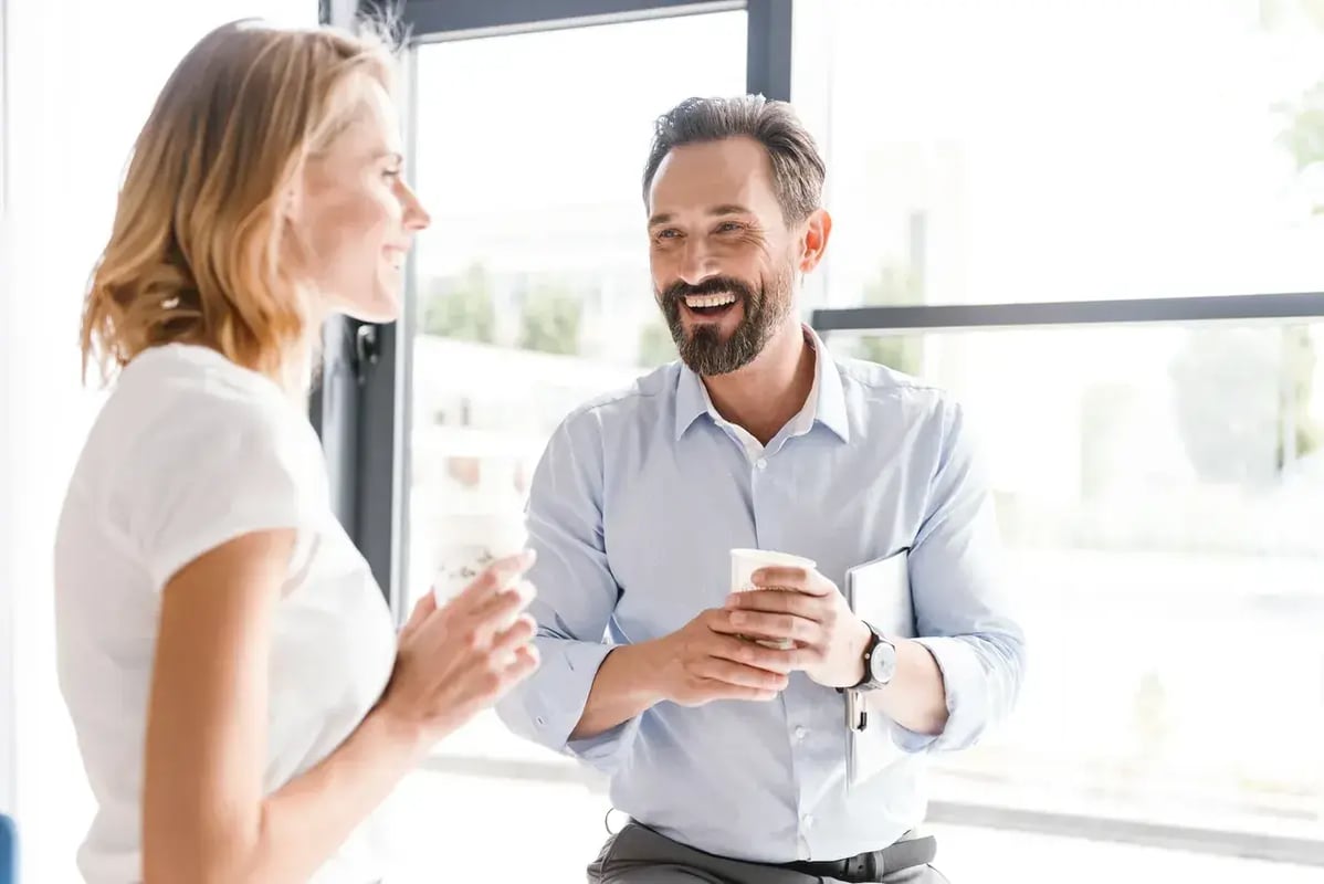 Twee collega’s lachen met een kop koffie bij het raam